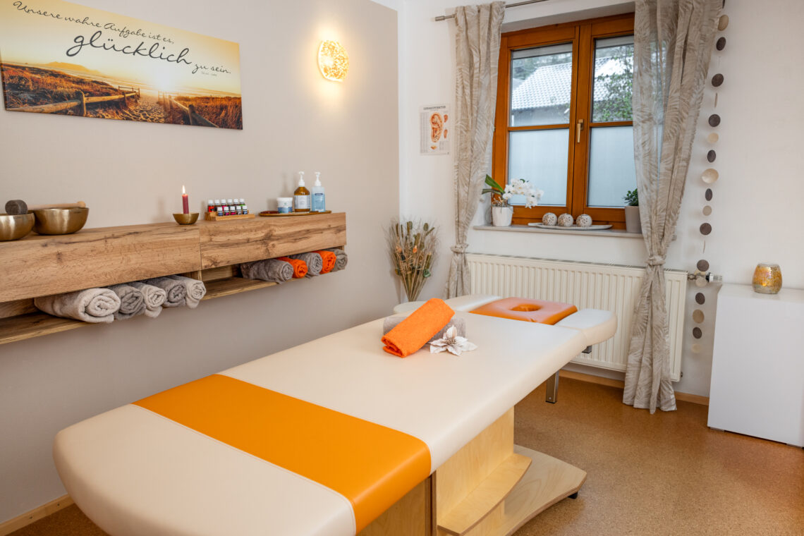 Massagepraxis Sarah Schedlberger. Es ist eine weiße Massageliege mit einem orangen Handtuch und einer Blume in der Mitte zu sehen. Hinter der liege sieht man ein Sideboard, dass mit Handtüchern befüllt ist und auf dem Massageöle stehen.