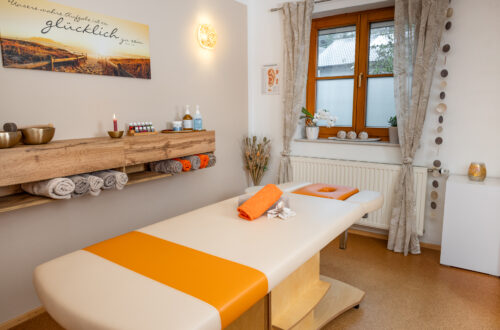 Massagepraxis Sarah Schedlberger. Es ist eine weiße Massageliege mit einem orangen Handtuch und einer Blume in der Mitte zu sehen. Hinter der liege sieht man ein Sideboard, dass mit Handtüchern befüllt ist und auf dem Massageöle stehen.